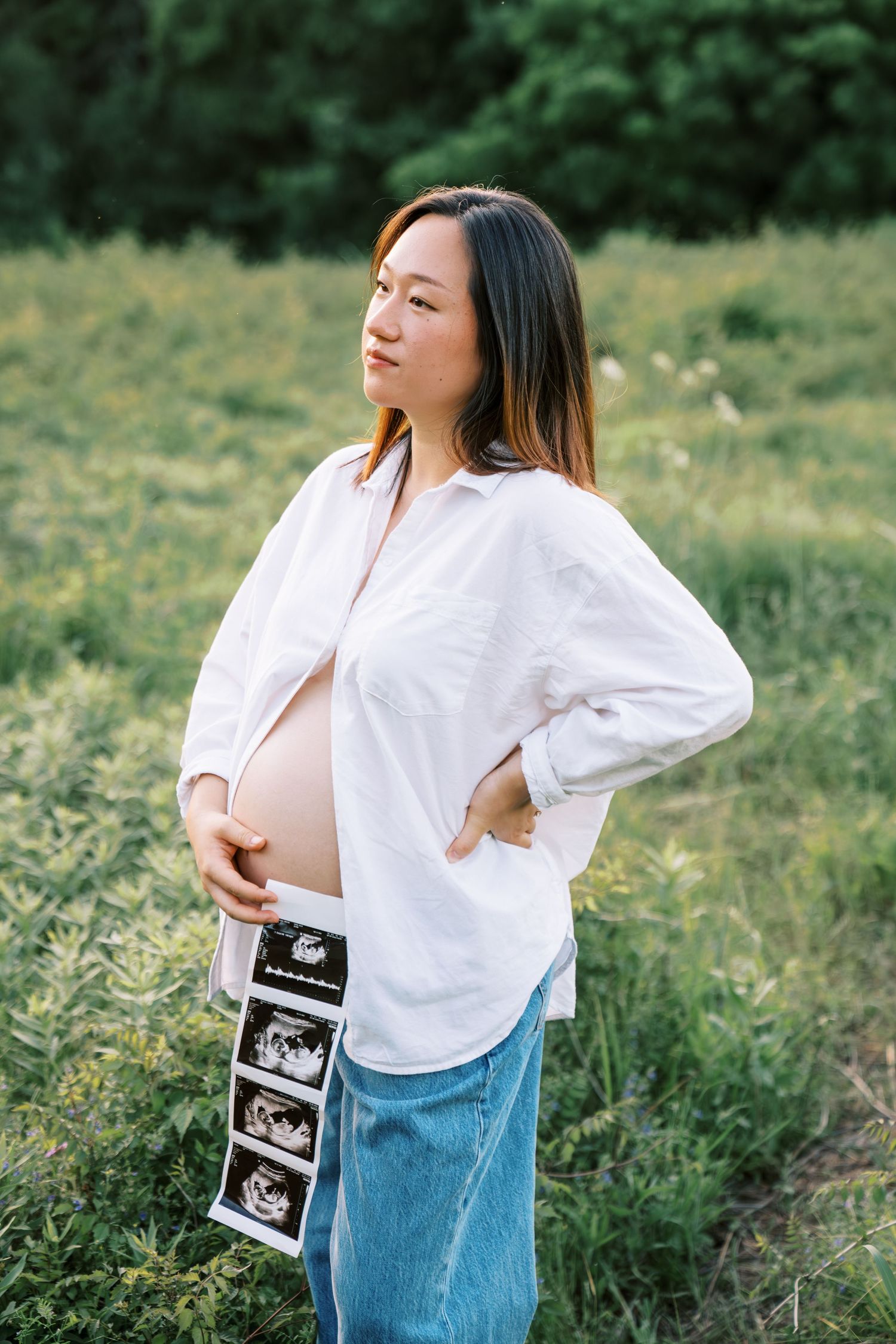 Pregnant Asian woman in white shirt and jeans holds ultrasound photos while standing in a green field.