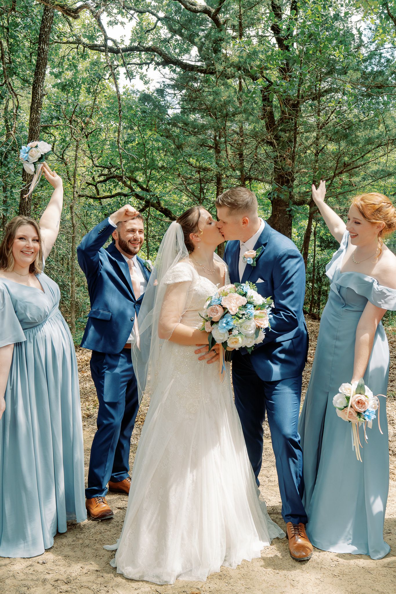 Wedding party celebrates joyfully on a wooded path with bridesmaids in light blue dresses and couple sharing a kiss.