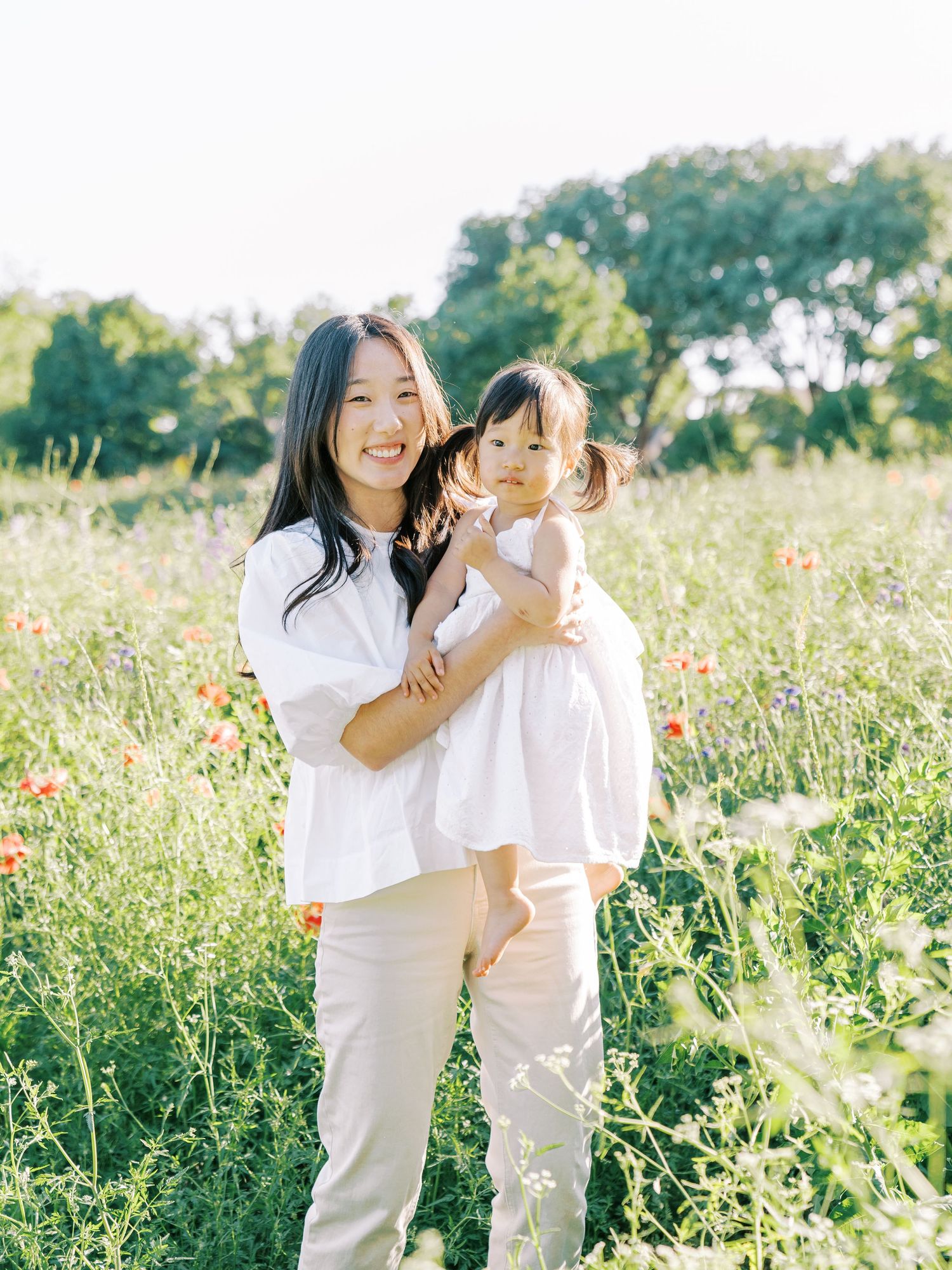 Mother and child wearing white outfits embrace while standing in a sunny wildflower meadow.