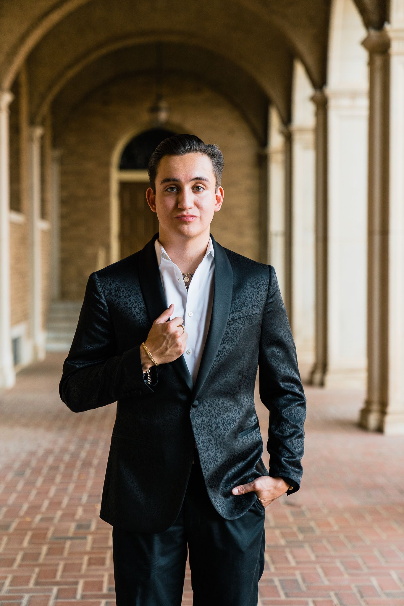 A person in a dark suit poses in an elegant arched hallway with brick flooring and classical architecture.