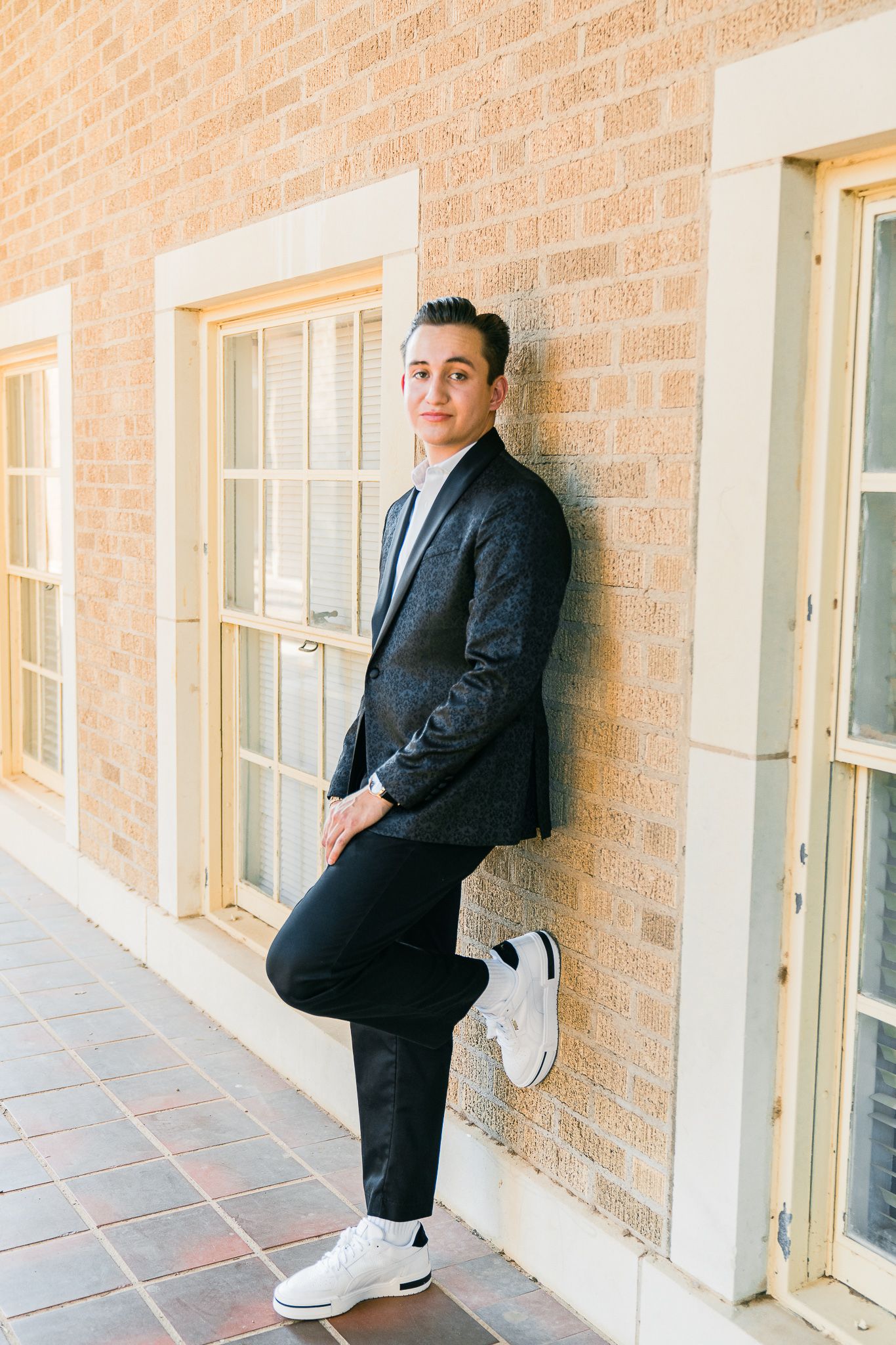 Person in dark suit and white sneakers poses against brick wall in three different casual stances.