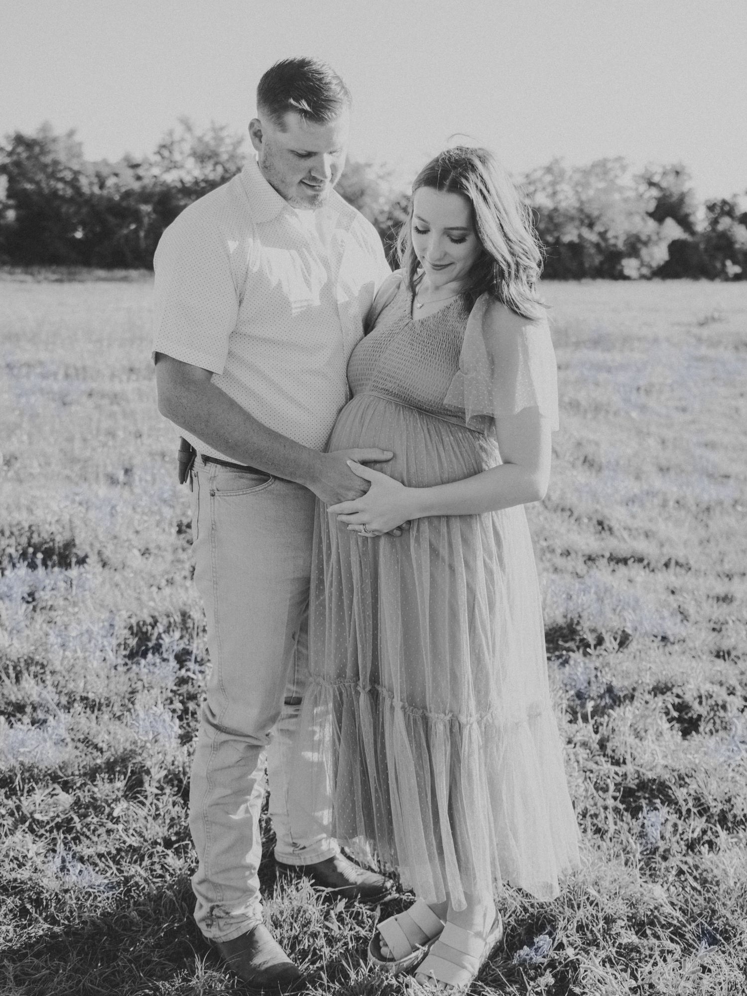 A black and white maternity photo captures a tender moment between a couple in a field.