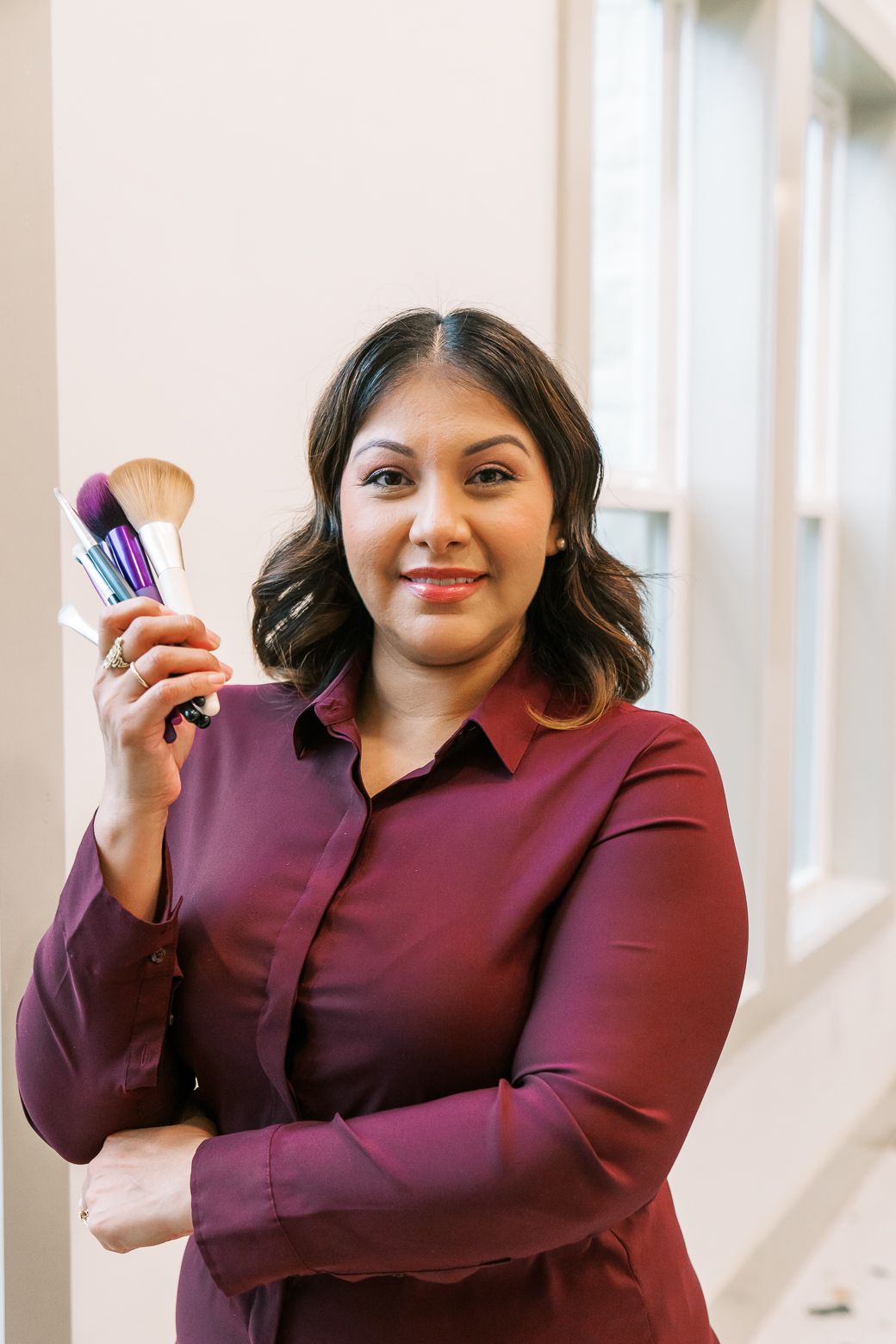 A person in a burgundy top displays makeup brushes while smiling in a naturally lit room.