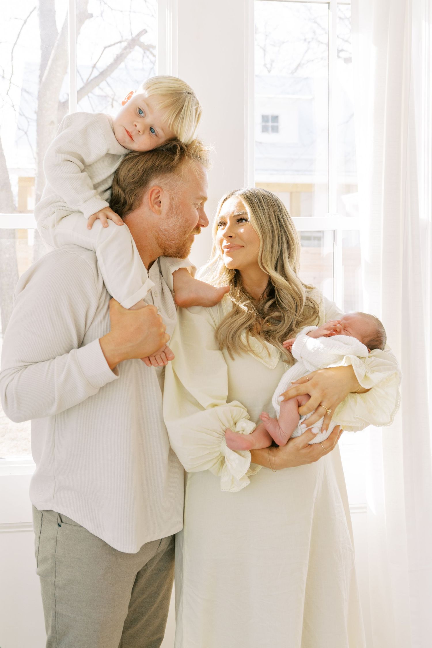 Family poses together by bright window in coordinated neutral outfits.