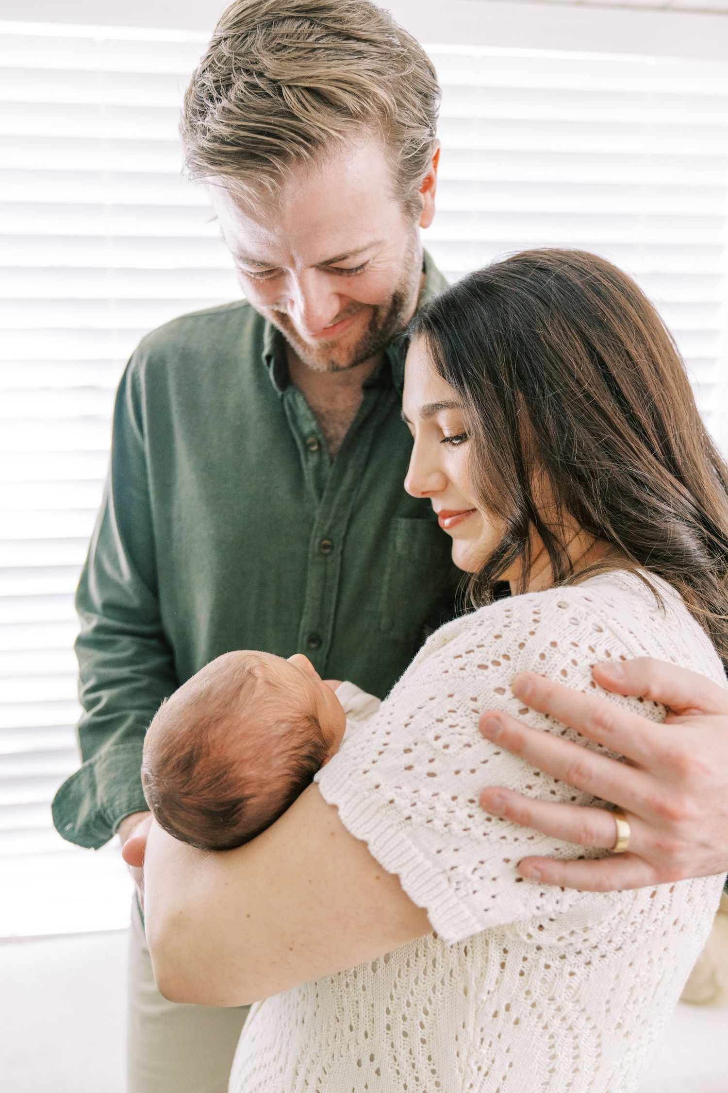 A couple in green and white sweaters share tender moments while holding their newborn baby in a bright room.
