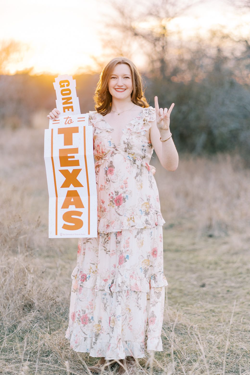Child wearing a floral dress holds white letter blocks outdoors during sunset in a grassy field.