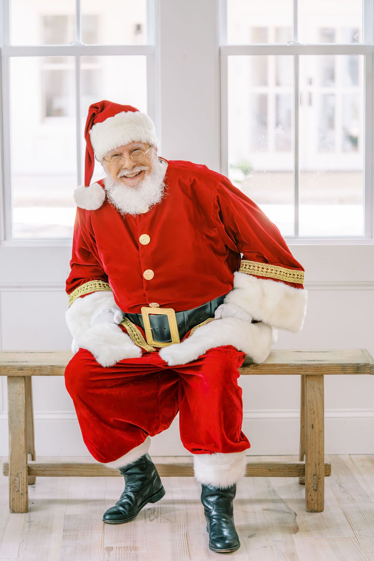 A jolly Santa Claus in red suit and black boots sits on a wooden bench, smiling warmly in a bright white room.