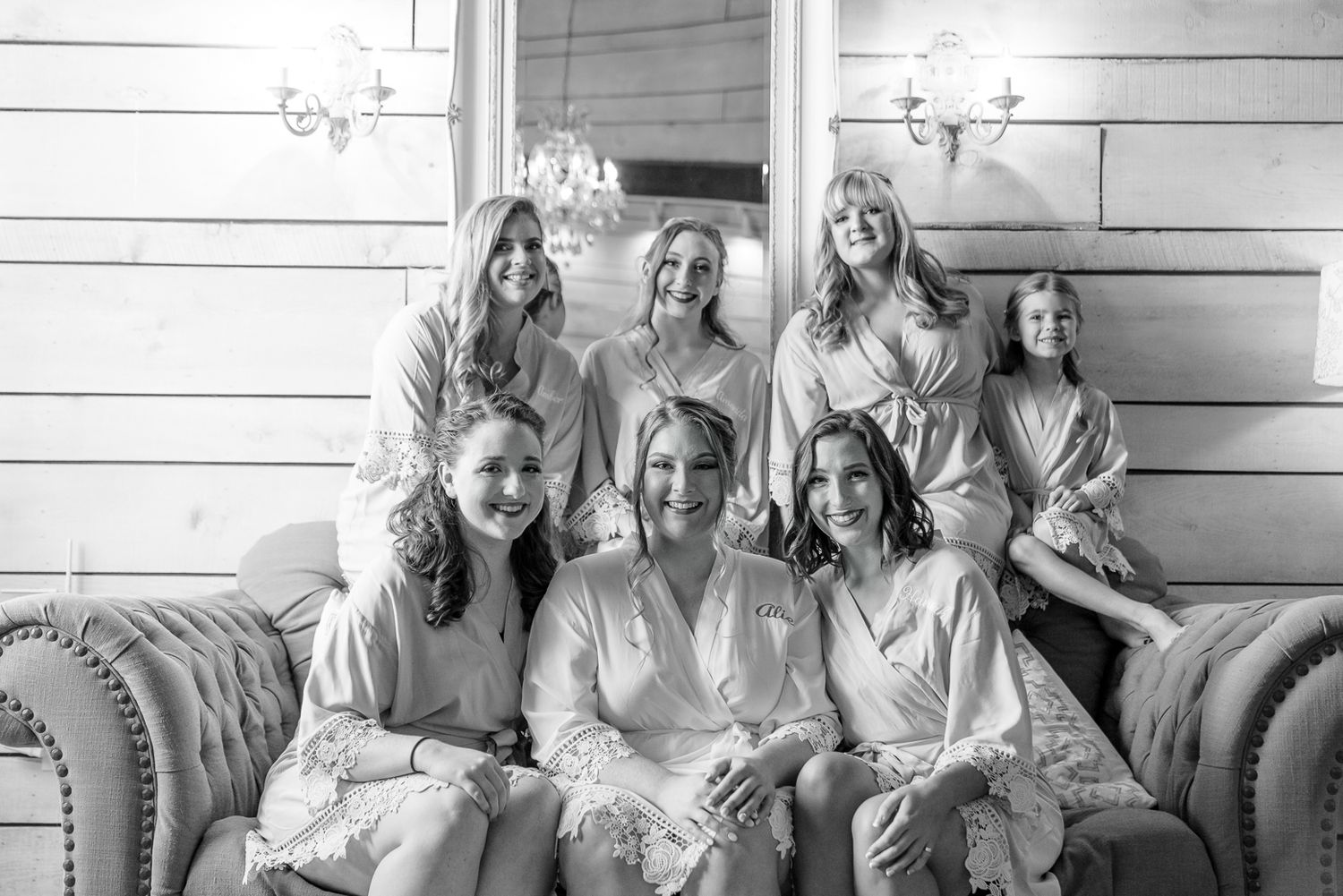Multiple people in coordinated white attire gather on a leather couch in a room with shiplap walls and sconces.