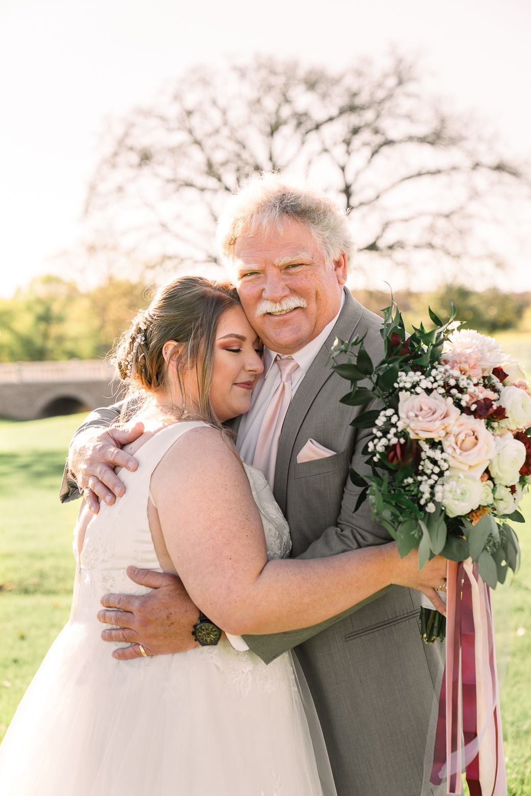 A tender wedding moment outdoors with a beautiful bouquet of pink roses and burgundy flowers against a sunny garden backdrop.