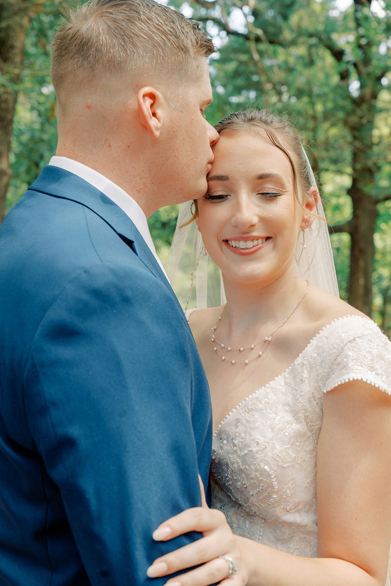 A tender wedding moment captured in a forest setting with the couple in navy suit and white lace dress.