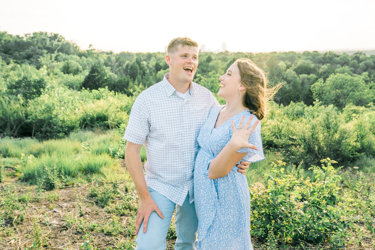 A couple in coordinating blue outfits laughs together while standing in a lush green field on a sunny day.