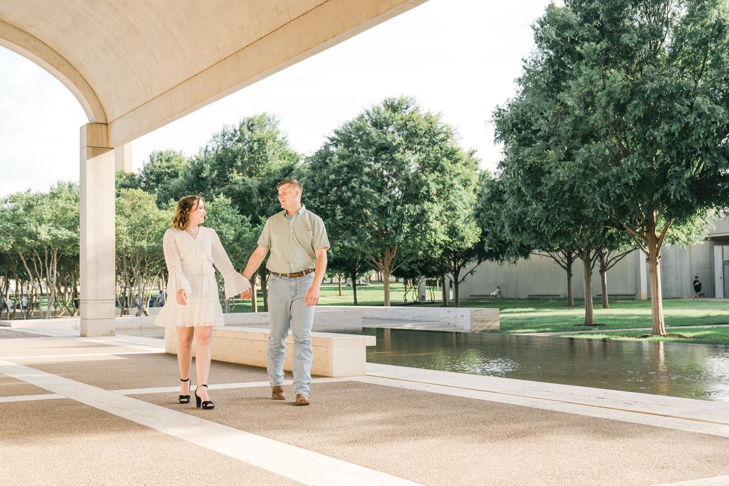A couple walks together under an arched walkway near a reflecting pool on a sunny day.