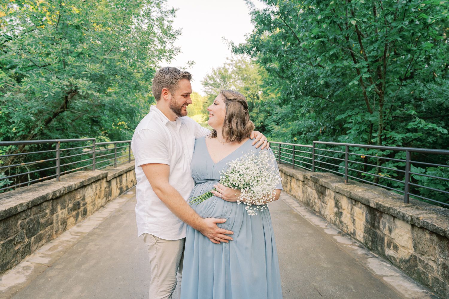 A romantic maternity photo on a stone bridge with the couple holding a bouquet of baby's breath flowers.