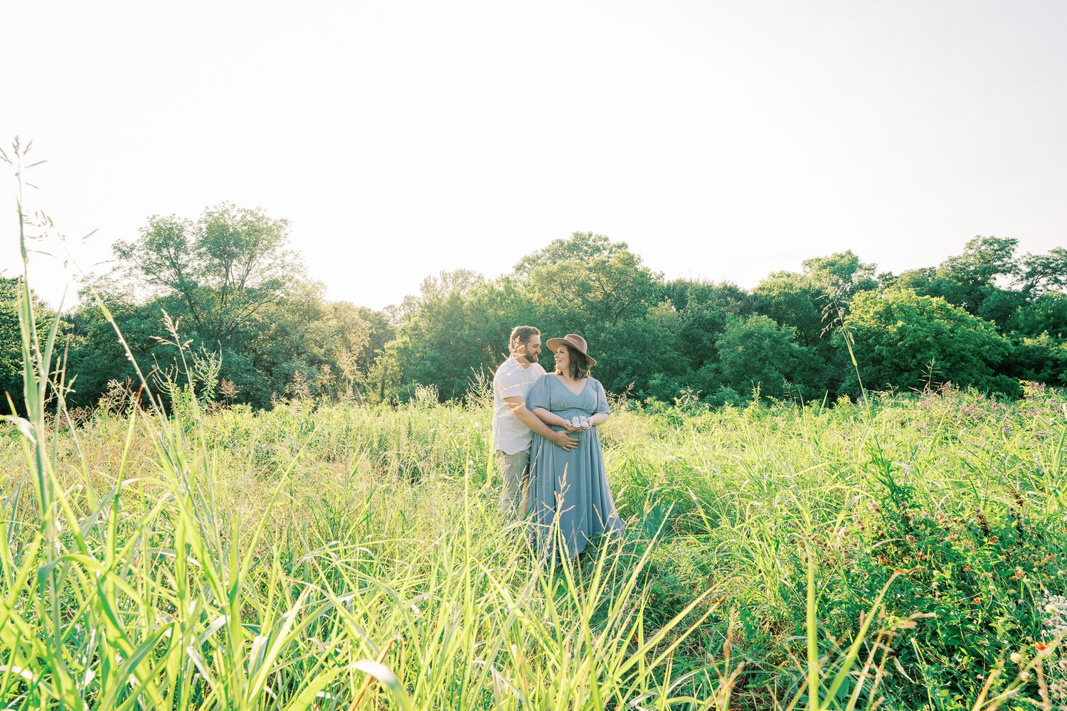 Couple walking together through a sunlit meadow with tall grass and wildflowers on a summer afternoon.