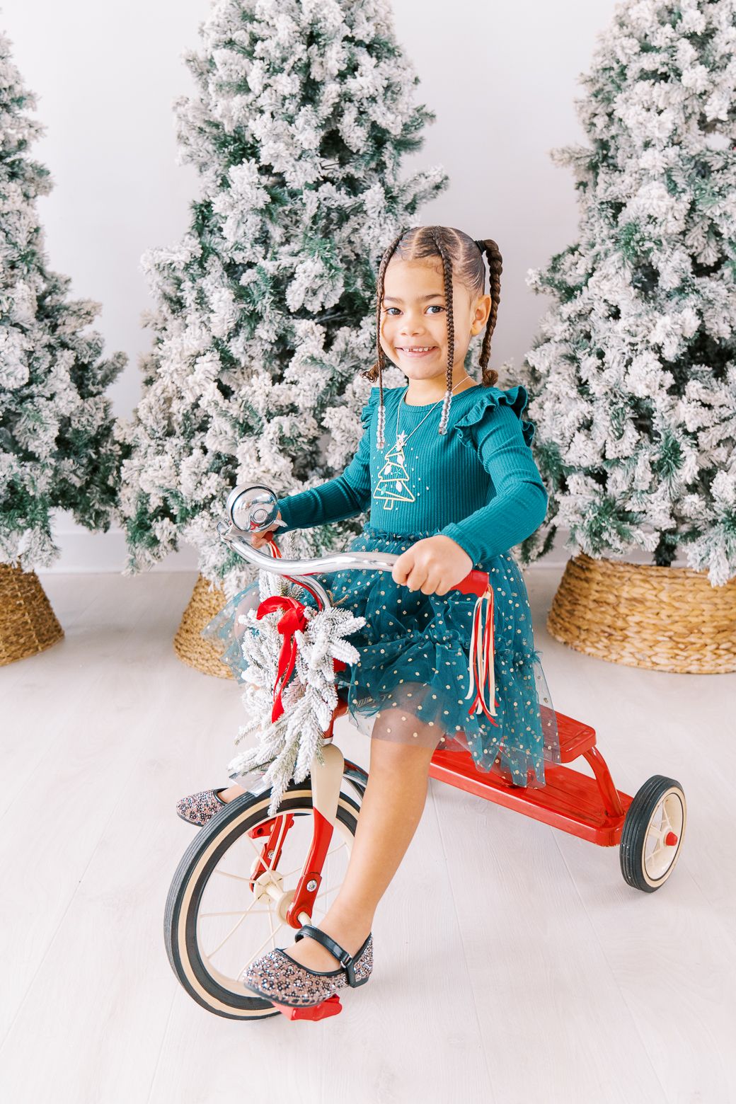 Child riding red tricycle in front of white flocked Christmas trees wearing teal holiday sweater.