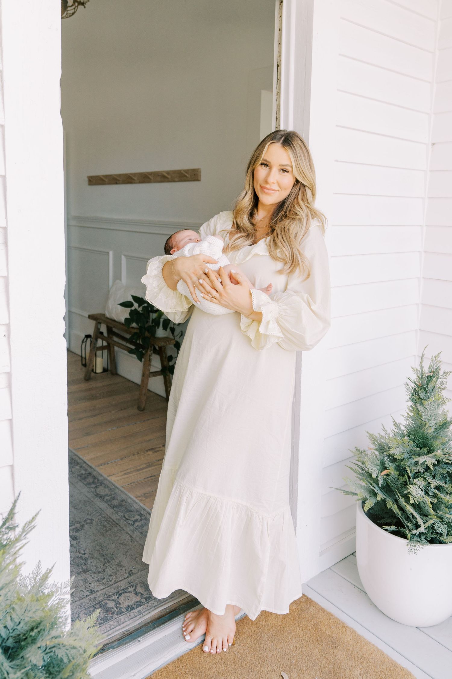 Woman in light dress holds baby near white pillars and decorative plants in modern home interior.