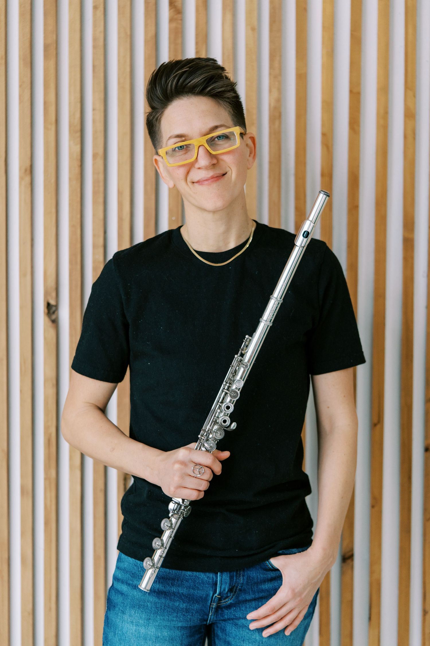 A musician in a black t-shirt and jeans poses with a silver flute against a striped wood panel wall.