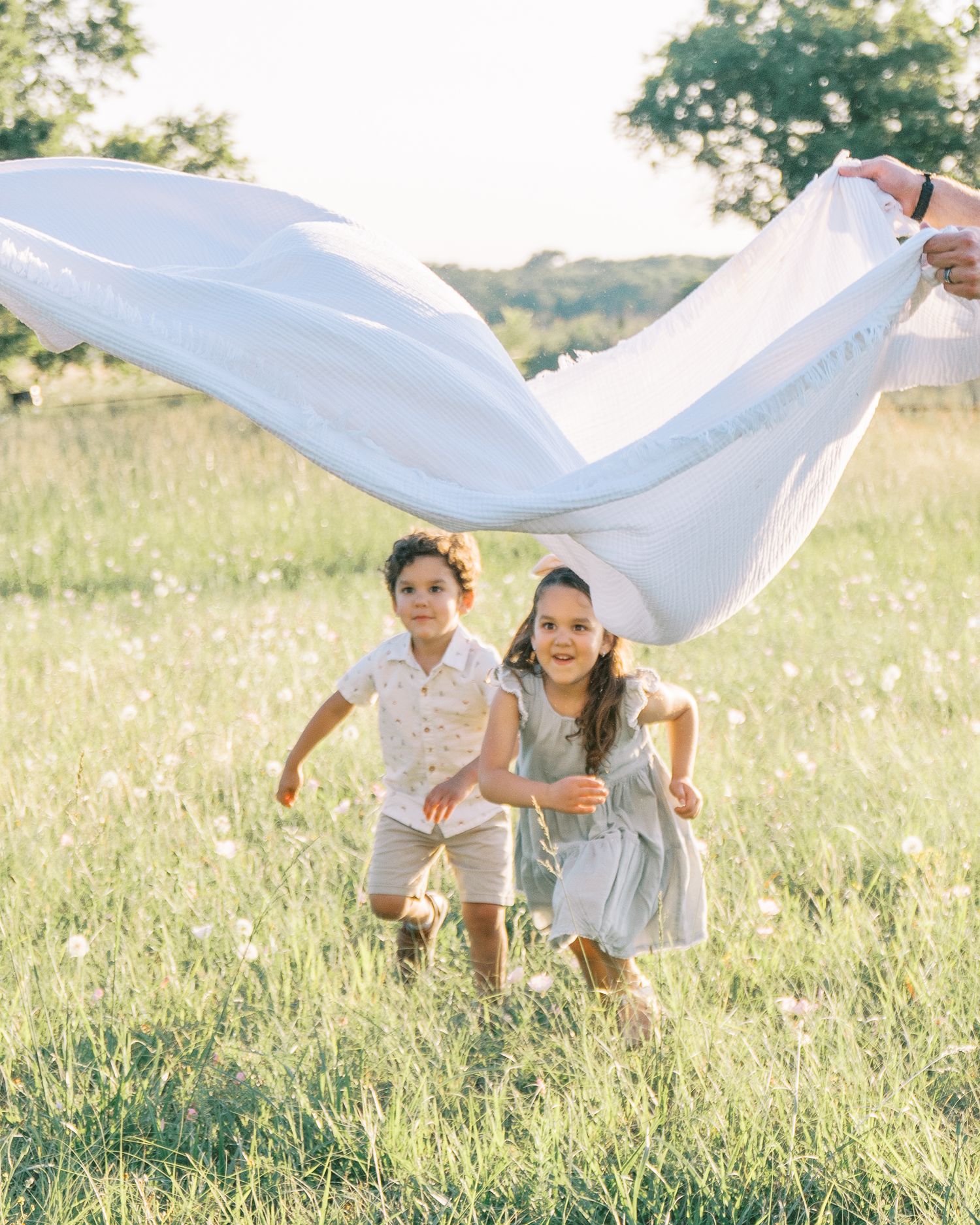 Two young children running joyfully through a sunny green meadow while an adult waves a white blanket above them.