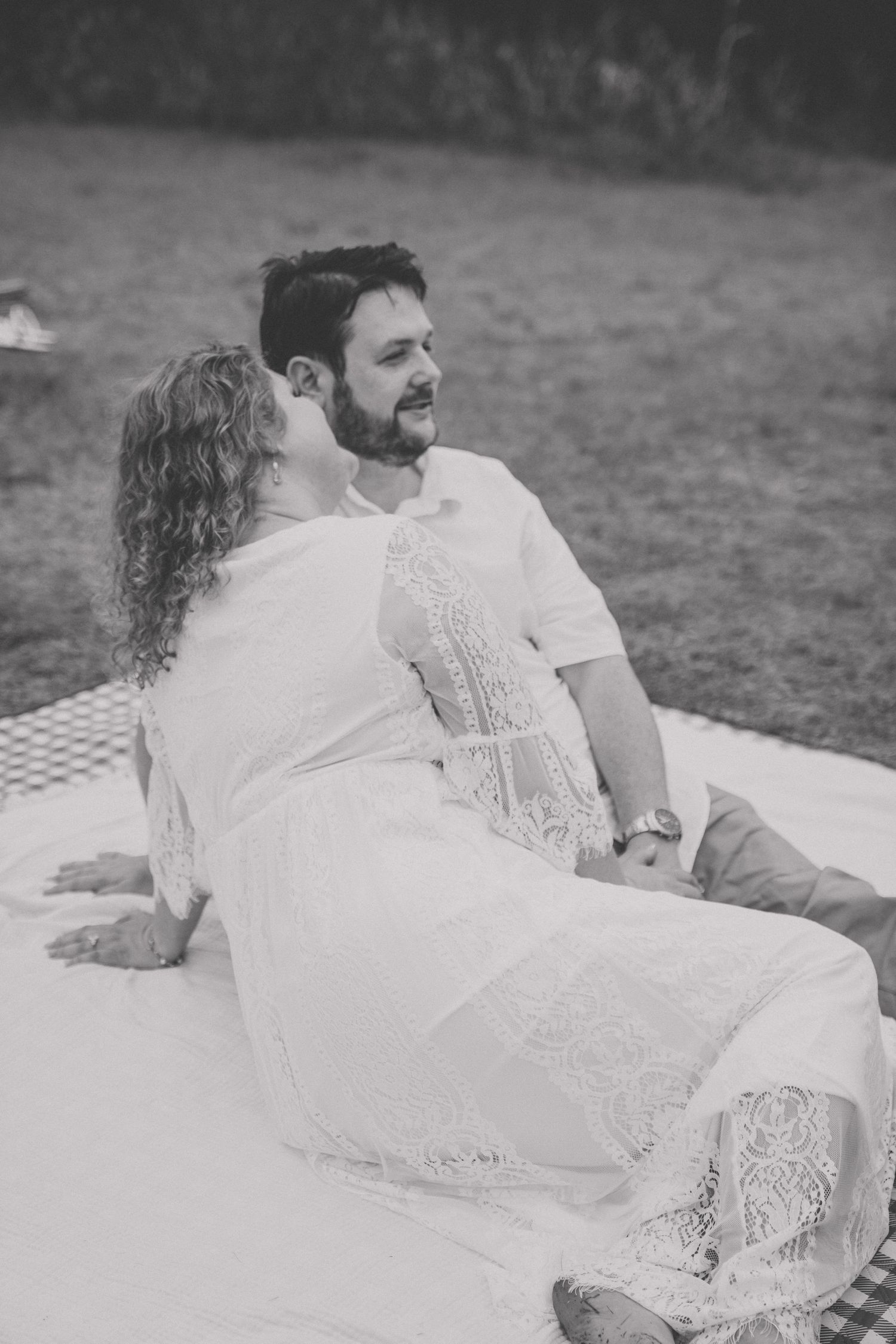 Black and white photo of a couple relaxing on a blanket outdoors, woman leaning in to kiss mans cheek.