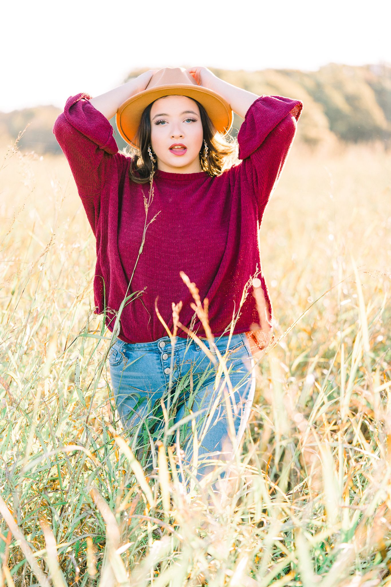 A casual autumn portrait in a burgundy sweater and tan hat standing in a golden wheat field.