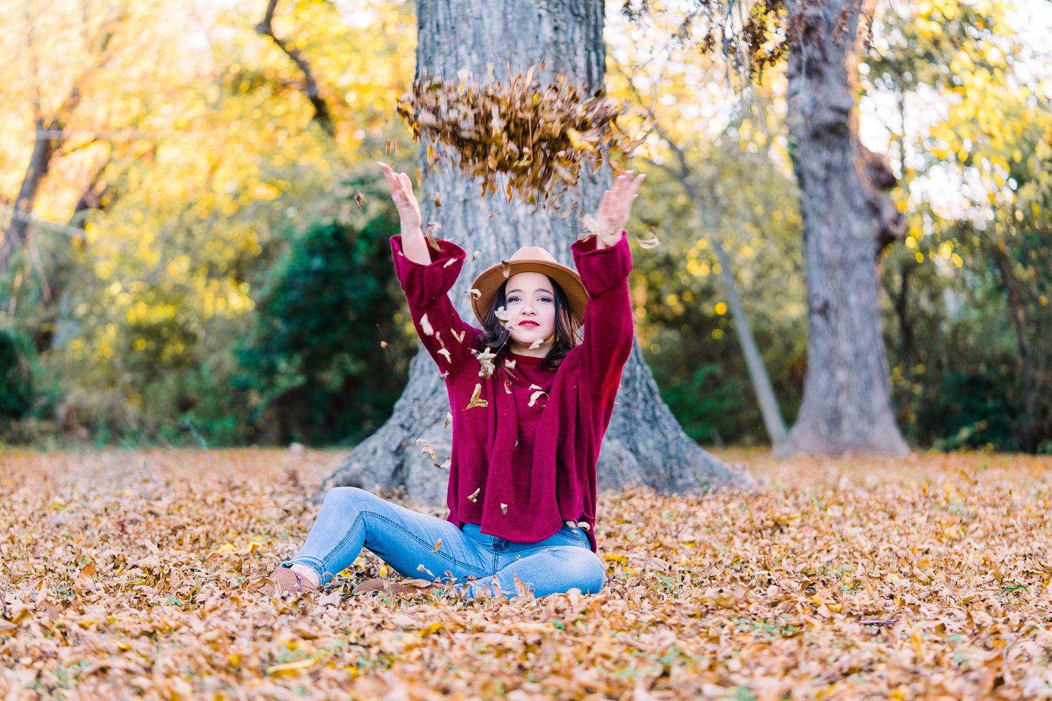 Person in a burgundy sweater and jeans playing in fallen autumn leaves on a sunny day.