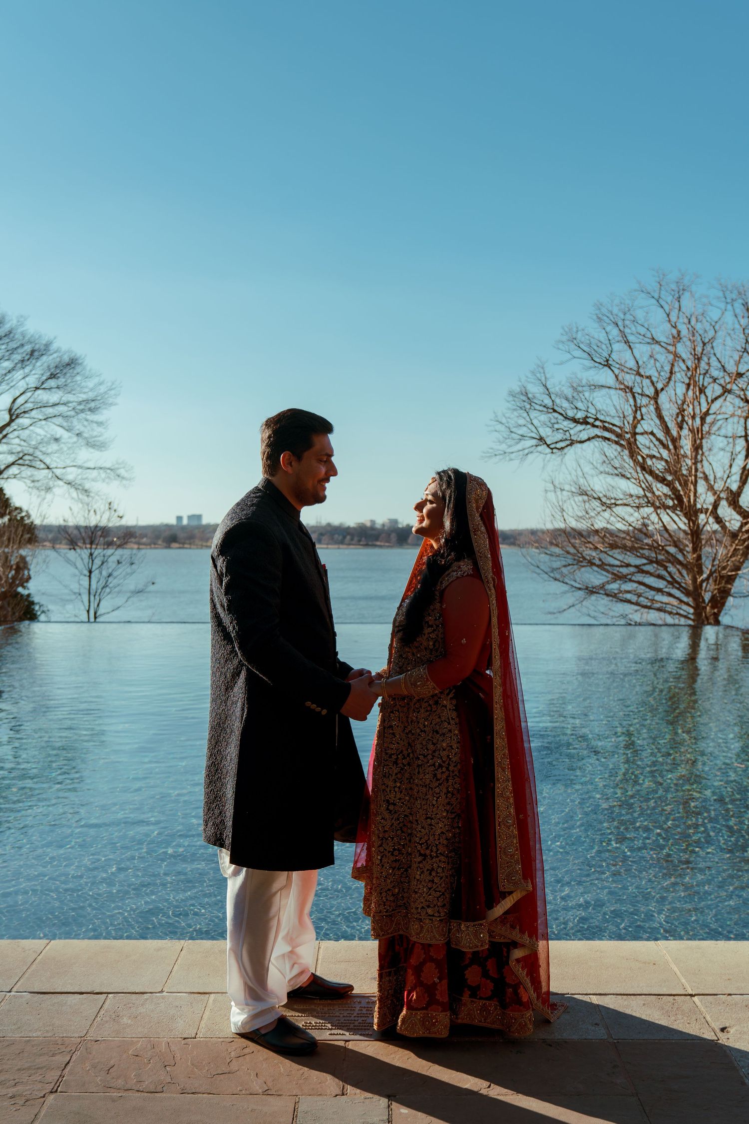 A couple in traditional South Asian wedding attire standing by a lake with bare trees in the background.