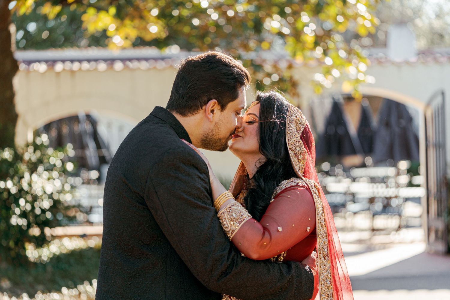 A couple shares a romantic kiss outdoors in traditional South Asian wedding attire with sunlit bokeh in the background.