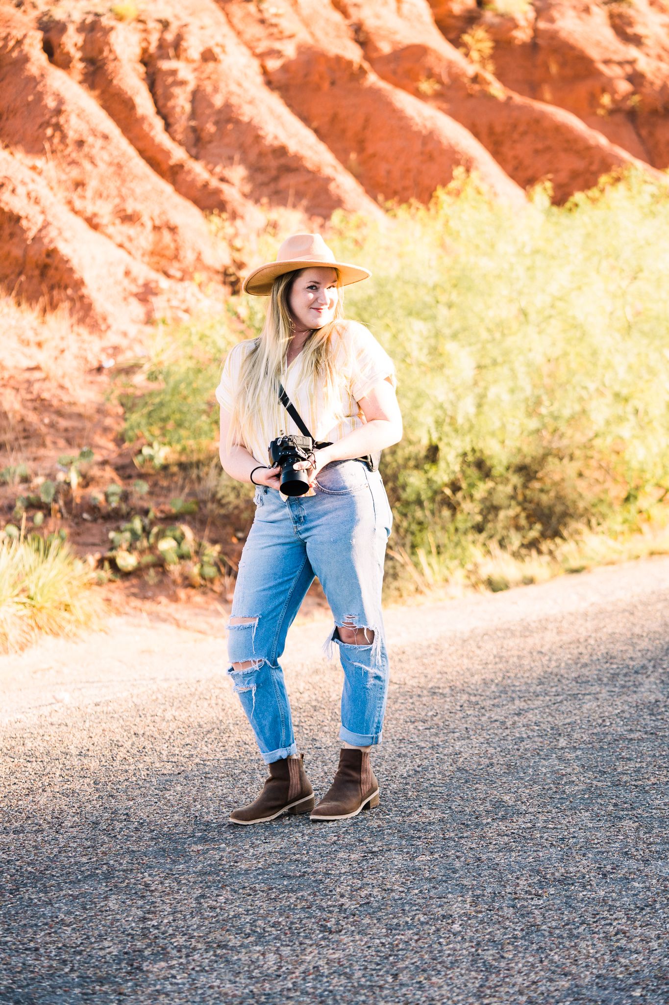 Person in cowboy hat and jeans kneels on asphalt road with red rock formations in background during sunset.