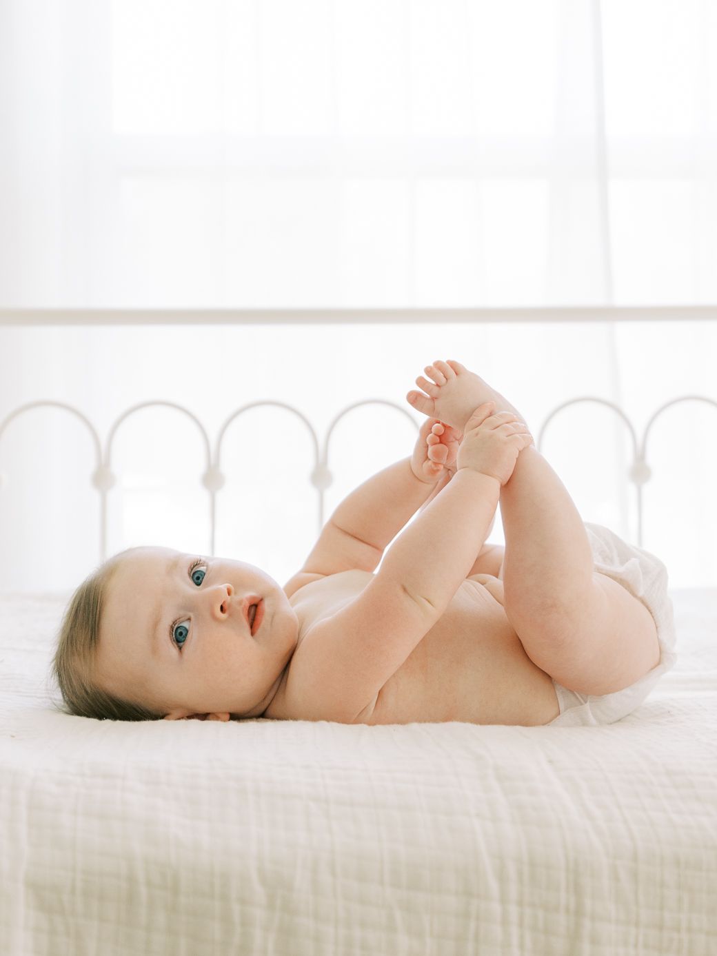 A baby lies on white bedding while looking up with bright blue eyes and chubby limbs.