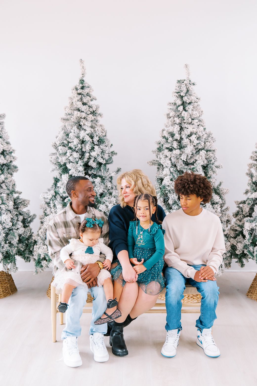 A group sits together on wooden chairs against a backdrop of frosted Christmas trees in a bright studio setting.