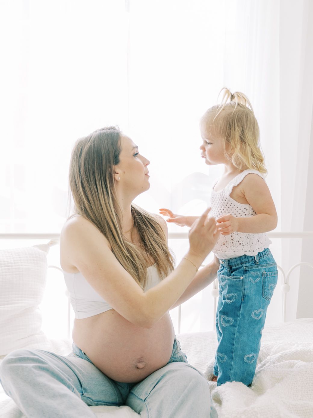 Intimate maternity photoshoot on white bedding showing playful moments between parent and toddler.