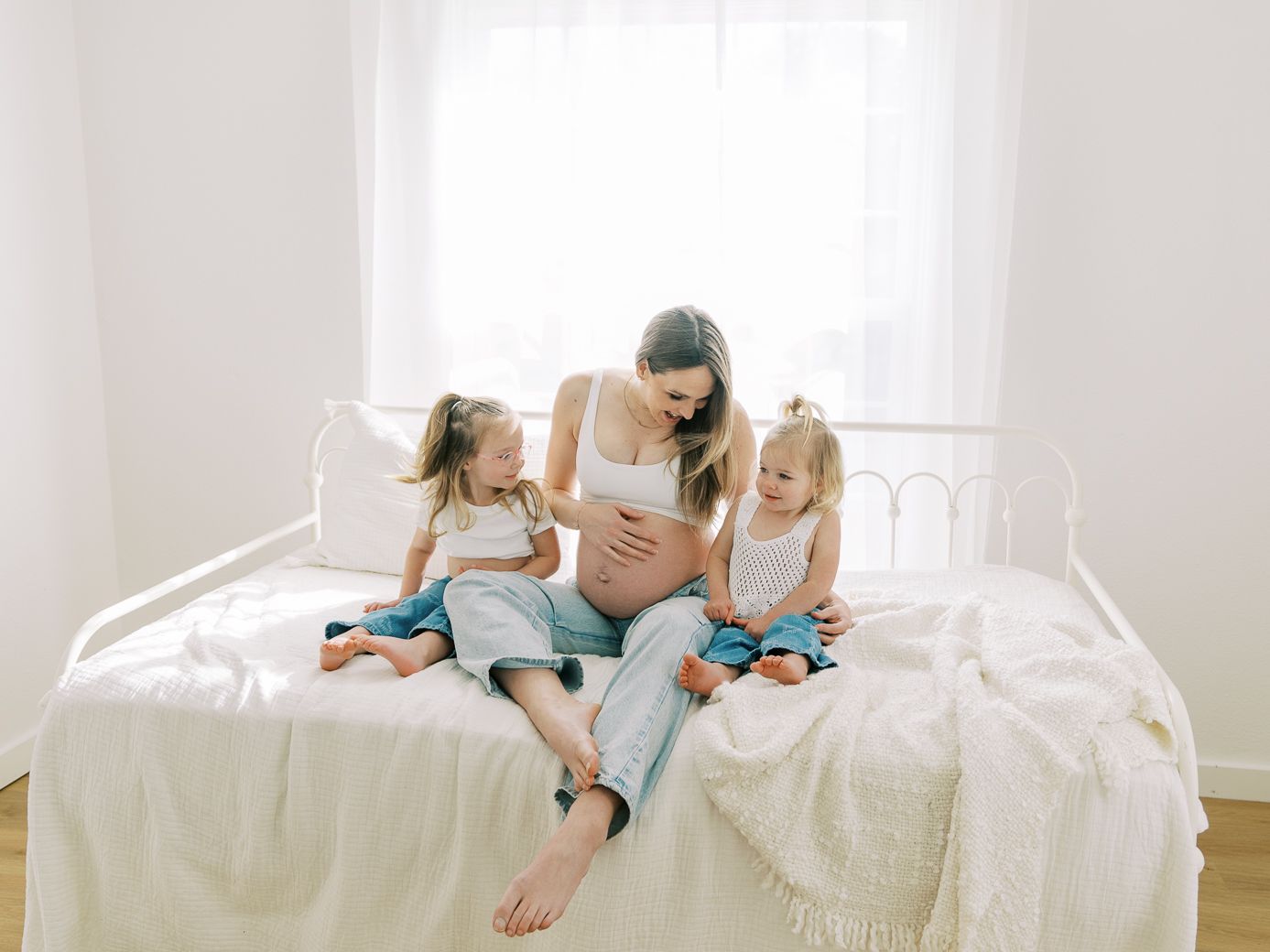 Lifestyle maternity session featuring relaxed poses on white bed with natural window lighting.