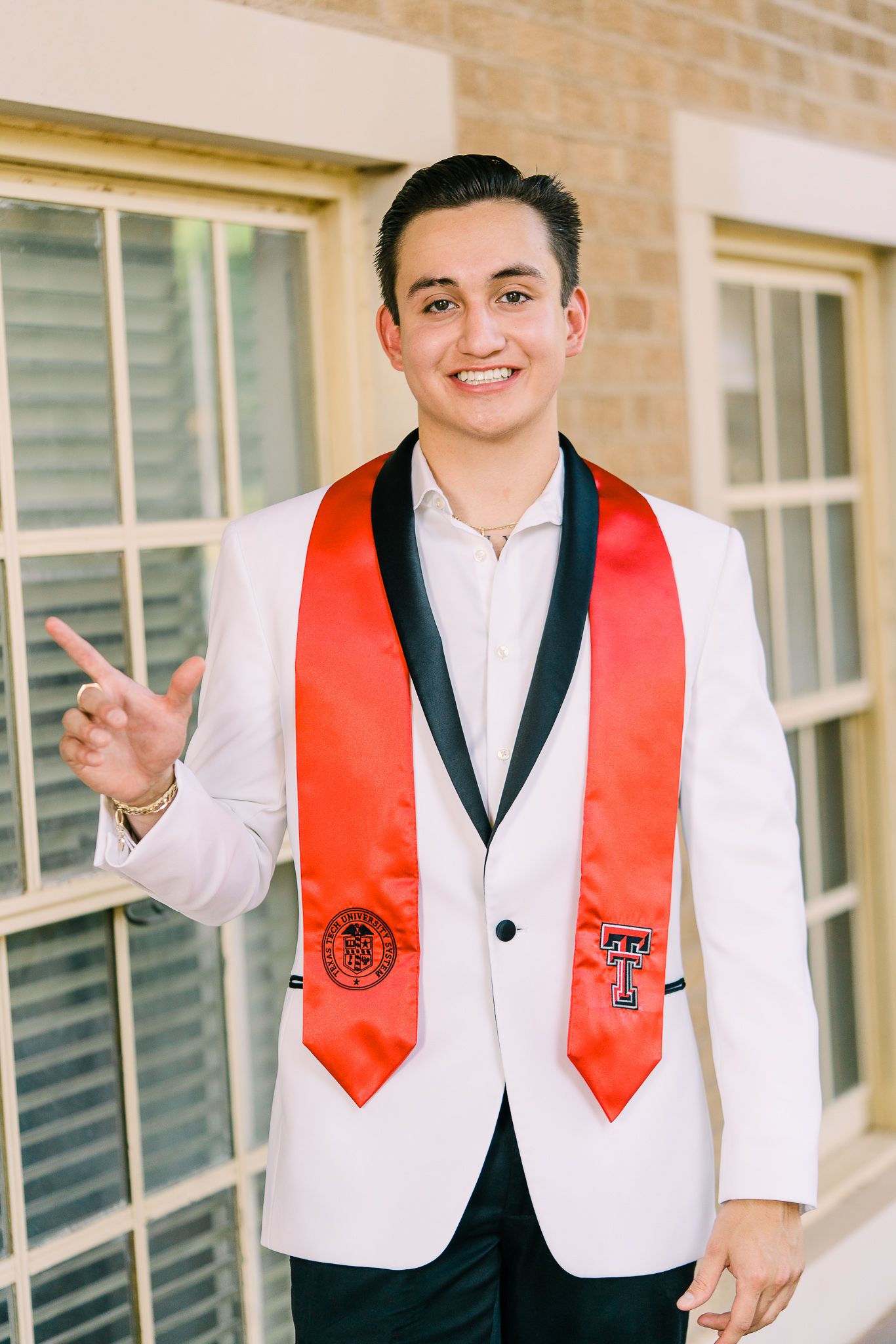 Graduate wearing white dress shirt and red graduation stole smiles while posing for photos outside brick building.