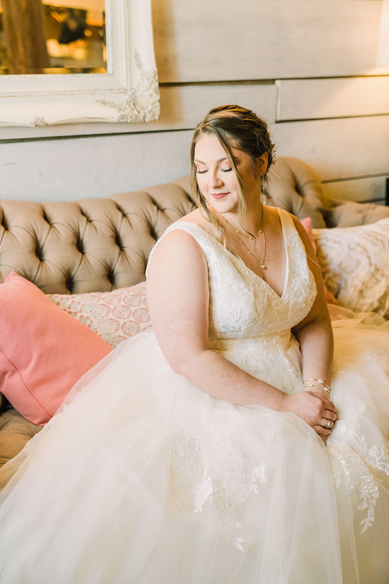 Bride in white lace gown reclining on tufted sofa with pink accent pillows.