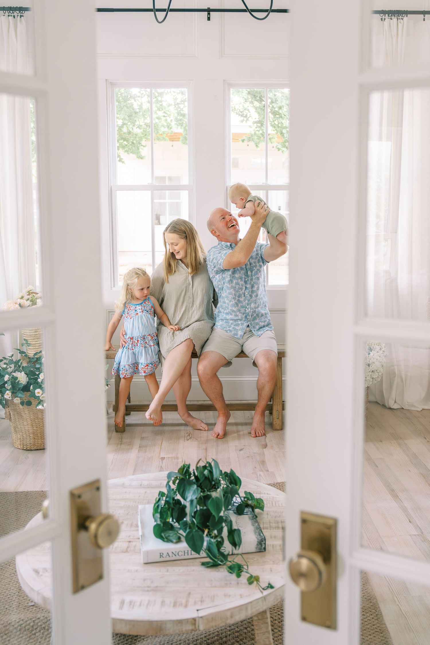 Happy family of four enjoying a bright, airy living room, with dad playfully lifting baby while mom and toddler daughter sit nearby.