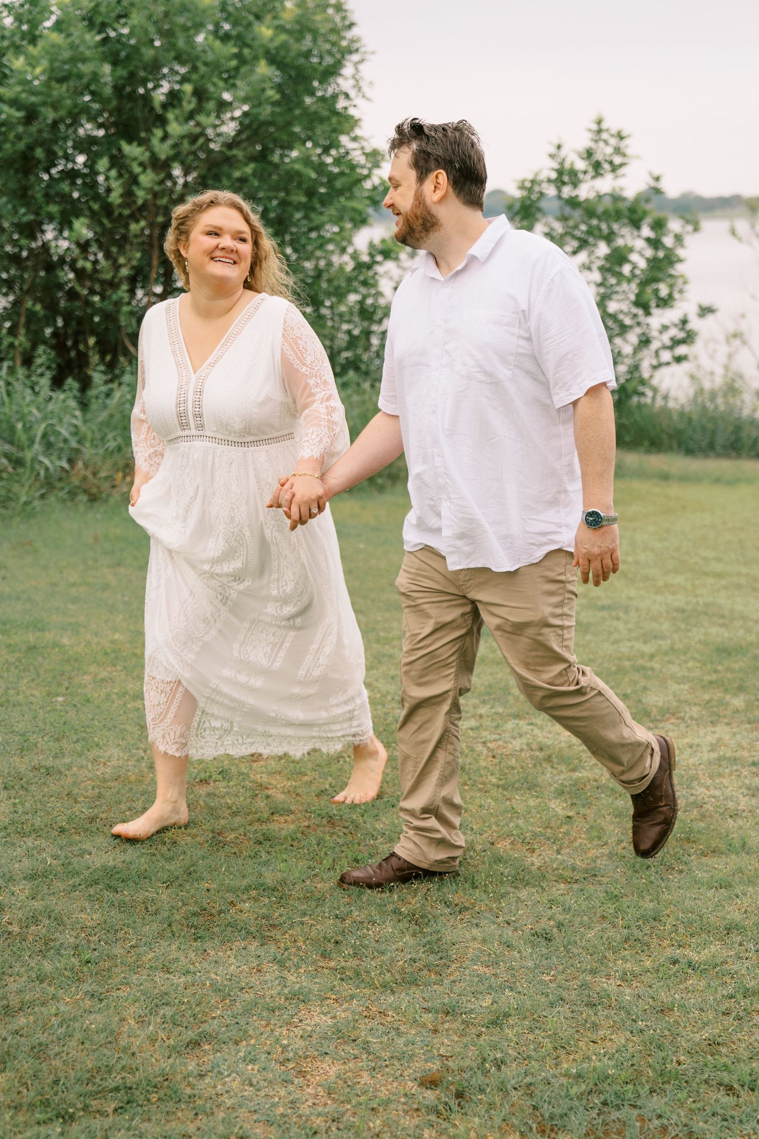 Happy couple holding hands and walking on green grass near a lake, woman in white lace dress, man in white shirt.