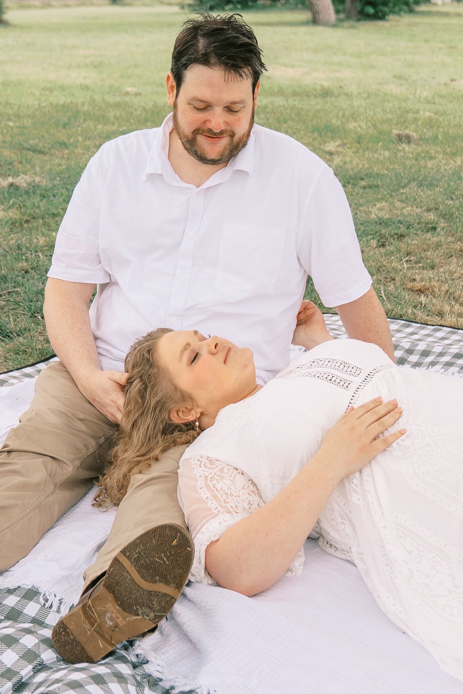 Pregnant woman in white lace dress lying on blanket with man in park during maternity photo session.
