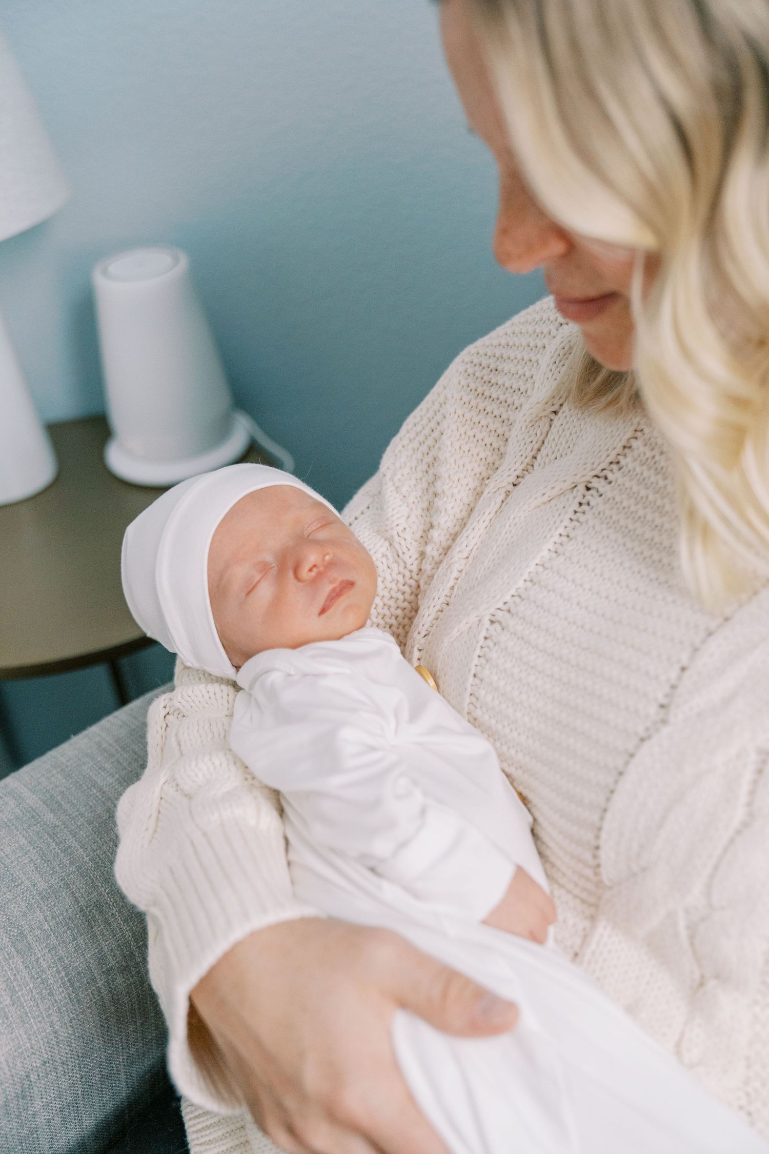 Newborn in white clothing being cradled in someone's arms wearing a cream knit sweater.