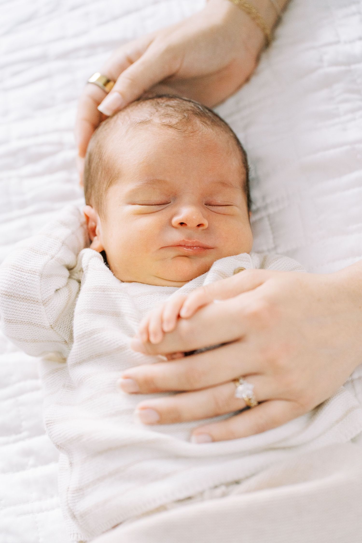 A gentle hand caresses a sleeping baby's face against soft white bedding.
