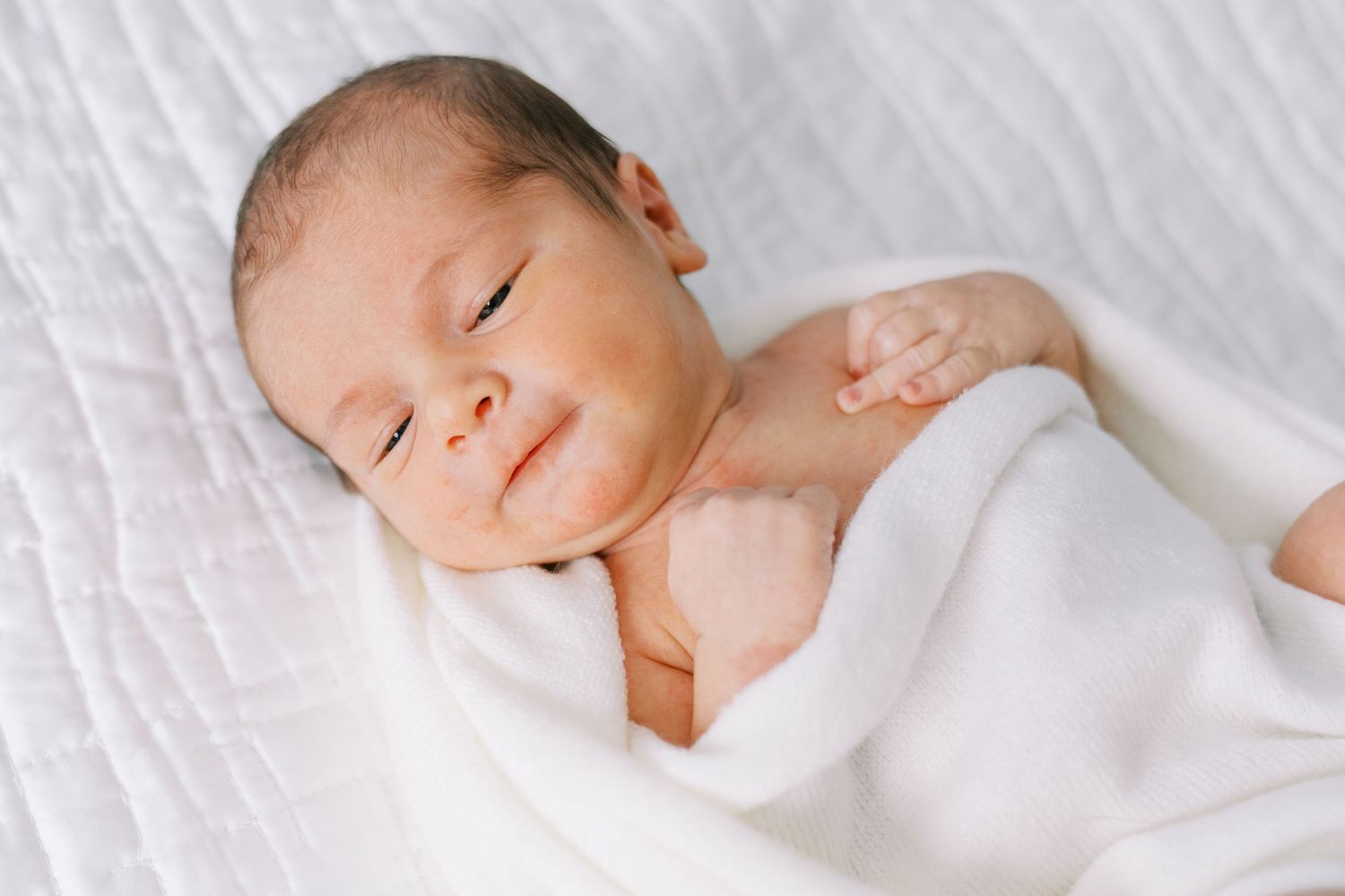 A peaceful newborn baby sleeping soundly on white bedding while wearing a soft white swaddle.