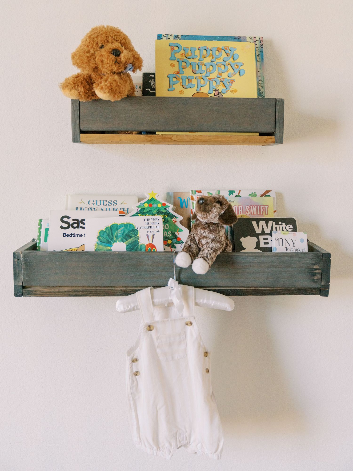 Two rustic gray wooden wall shelves displaying children's books, stuffed animals, and a white baby romper hanging below.