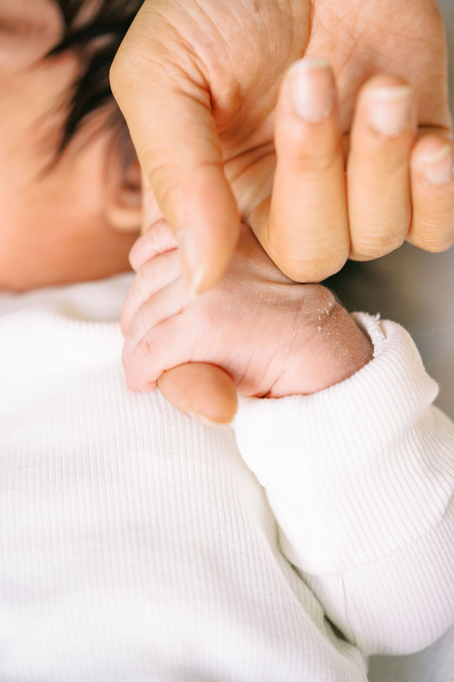 A newborn baby grasping an adult's finger with tiny hand, wearing white onesie, close-up bonding moment.