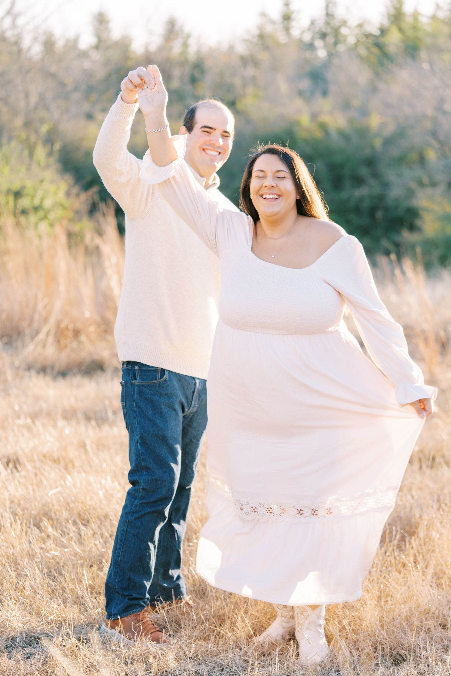 Joyful couple dances in field wearing coordinated white outfits during sunset.