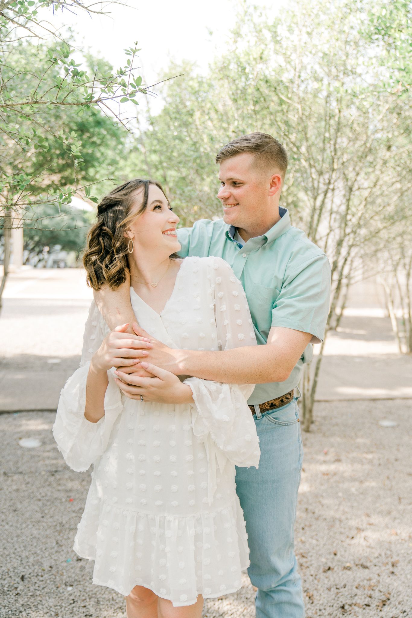 Couple embraces and kisses during an outdoor photo session among trees in soft natural lighting.