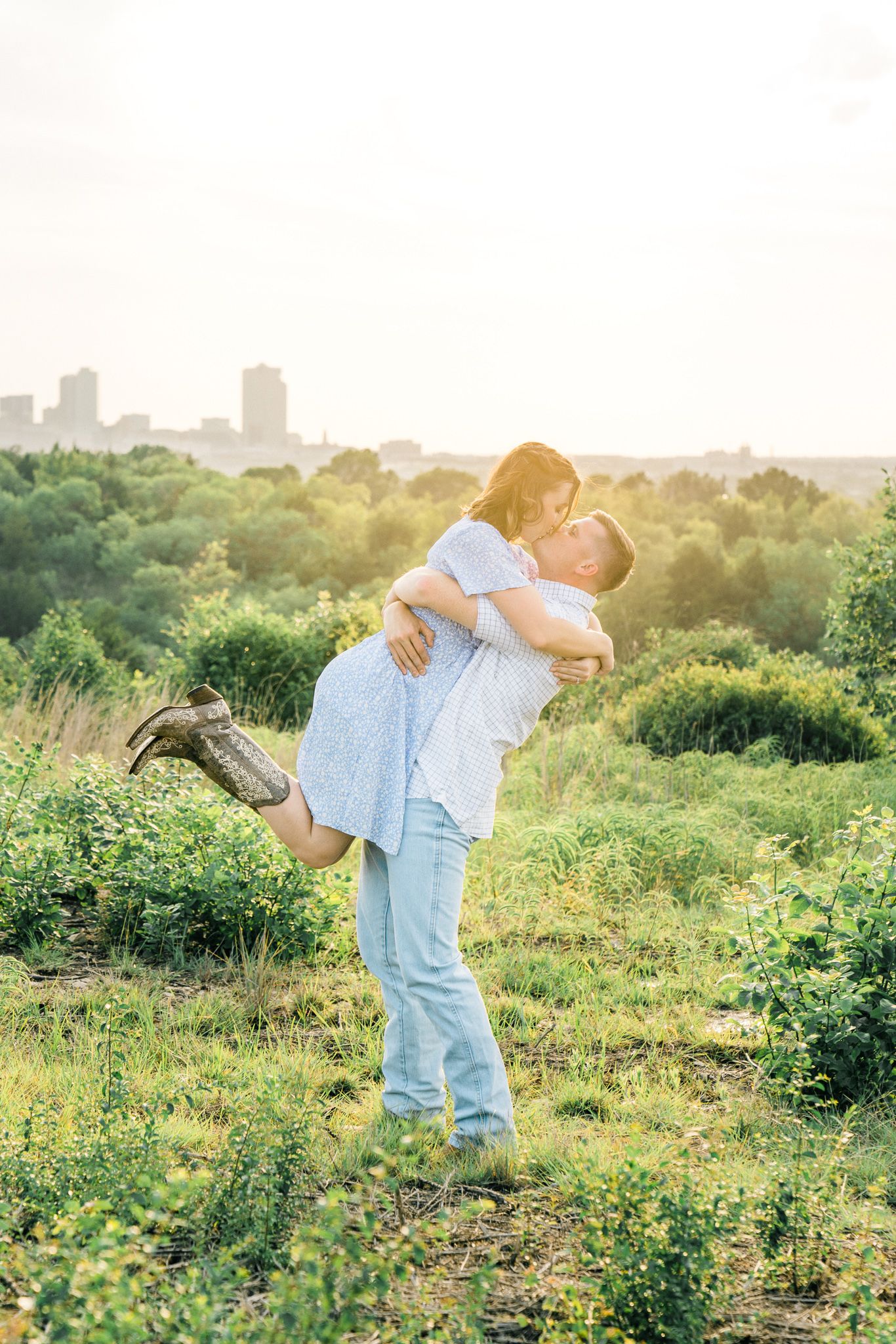 A couple dances playfully in a sunlit meadow wearing casual blue outfits against a city skyline backdrop.