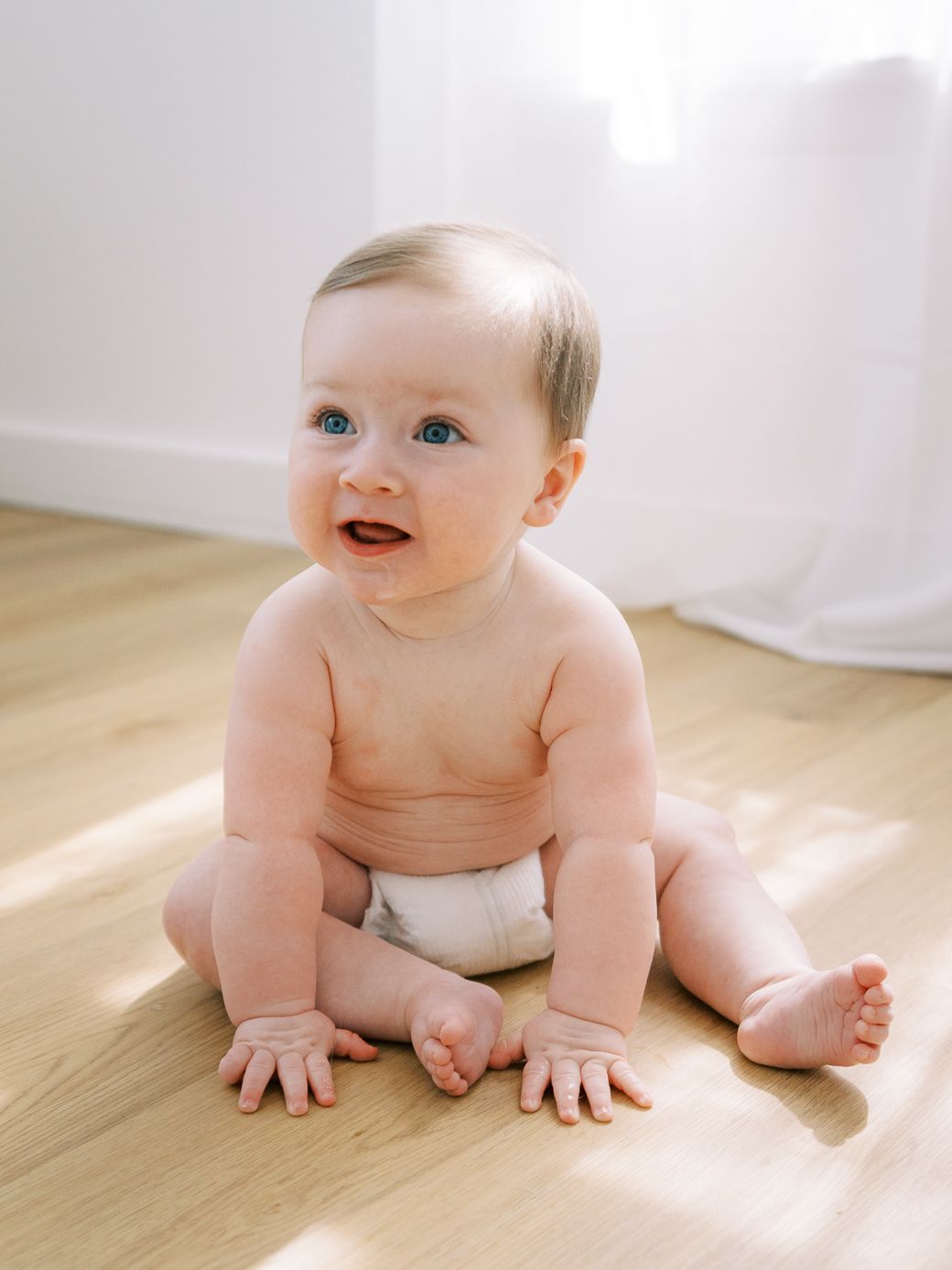 Baby in diaper crawls on wooden floor in natural light from window.