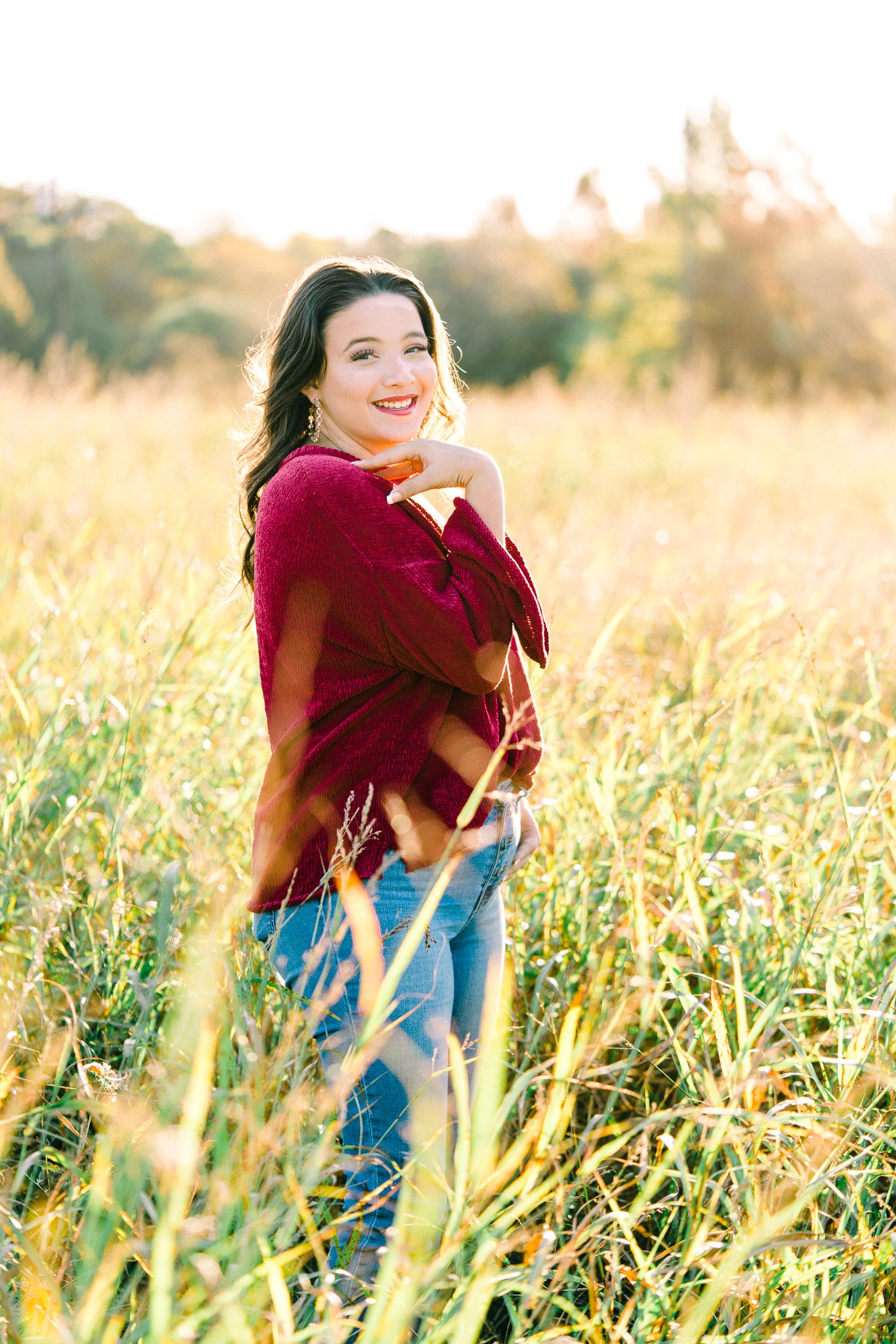 Person in red top and jeans smiles while posing in golden grass during magic hour.