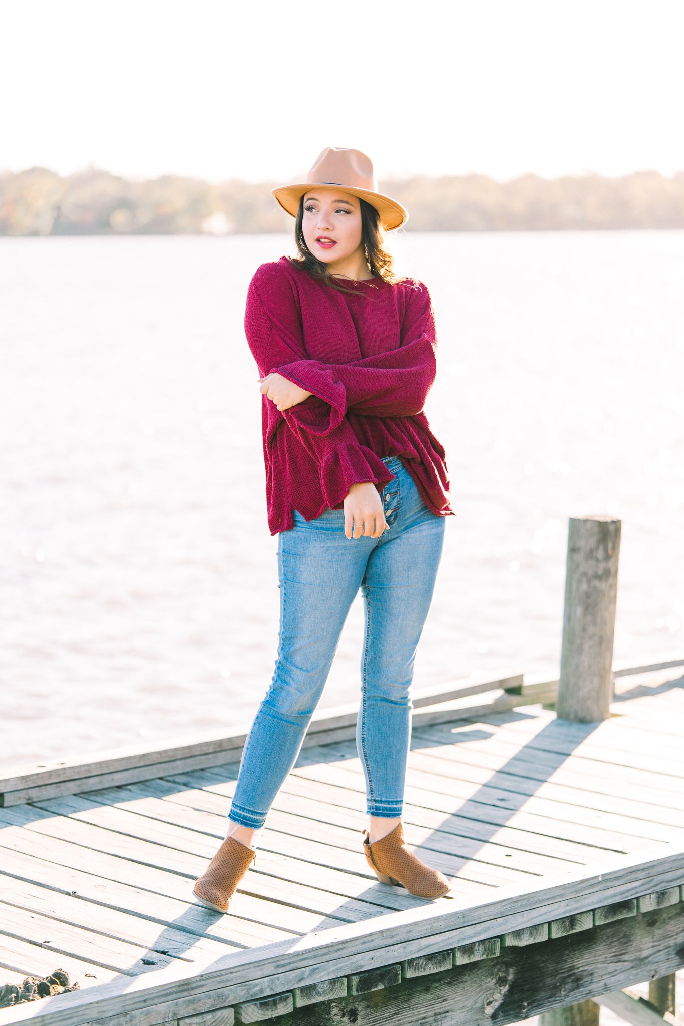Person in burgundy sweater and jeans poses playfully on wooden dock at sunset wearing tan wide-brim hat.