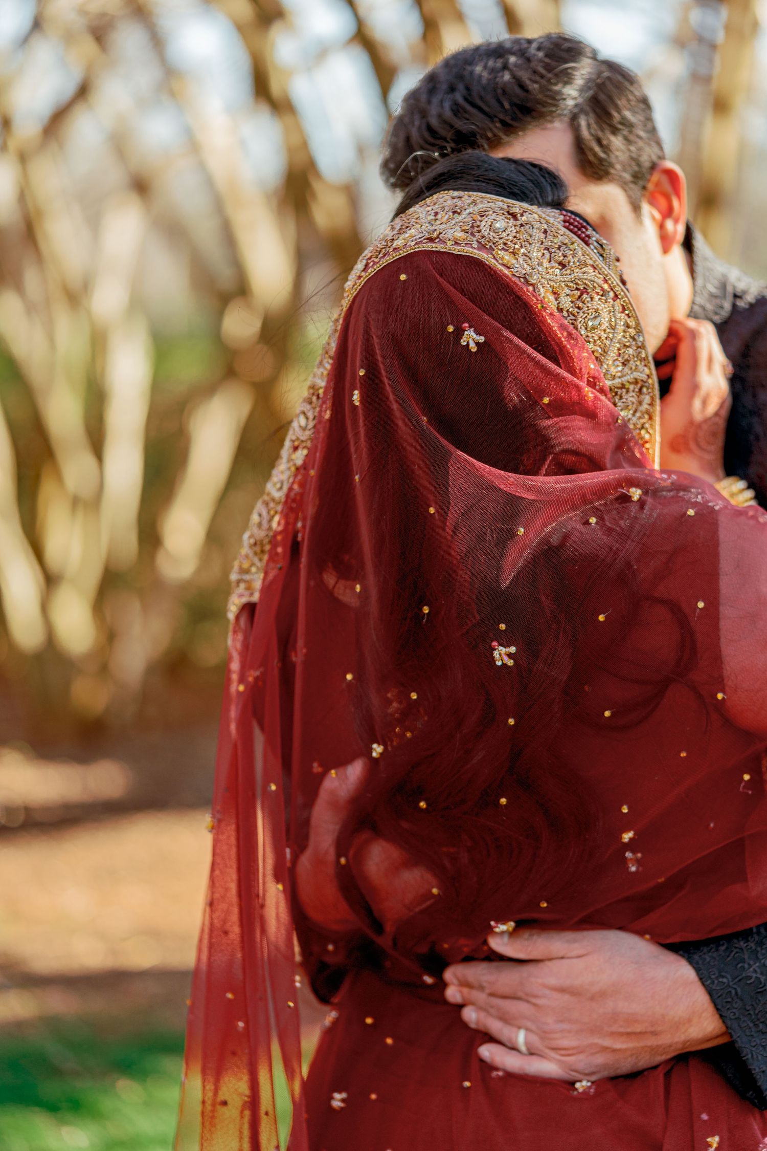 Couple embracing in burgundy traditional attire with golden details in an outdoor setting.