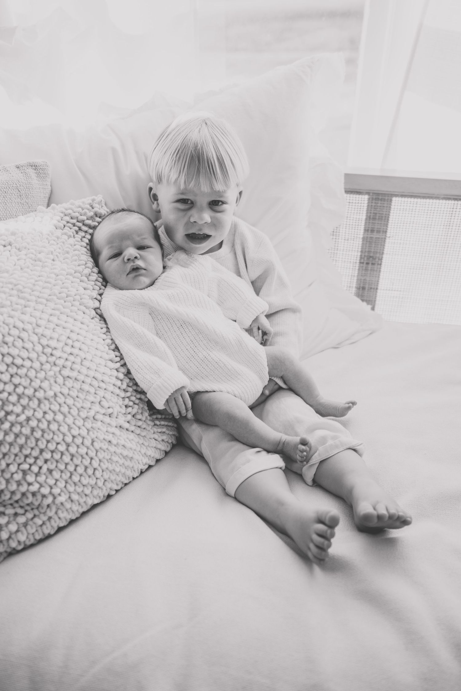 Two young siblings in white clothing resting on a white textured bed.